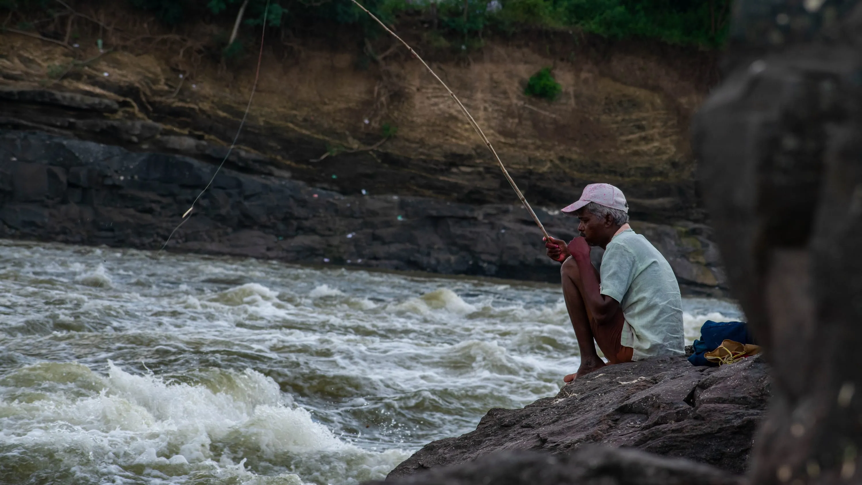 A man crouched on a rock casting a line into white water running fast between boulders.