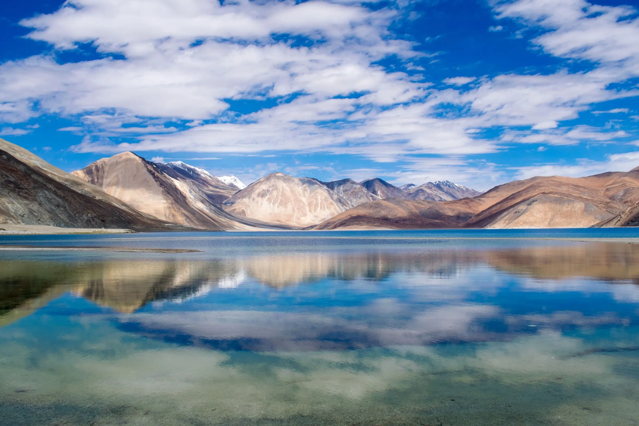 A still lake mirroring a bank of cloud and a low ridge, snow-lined mountains behind.