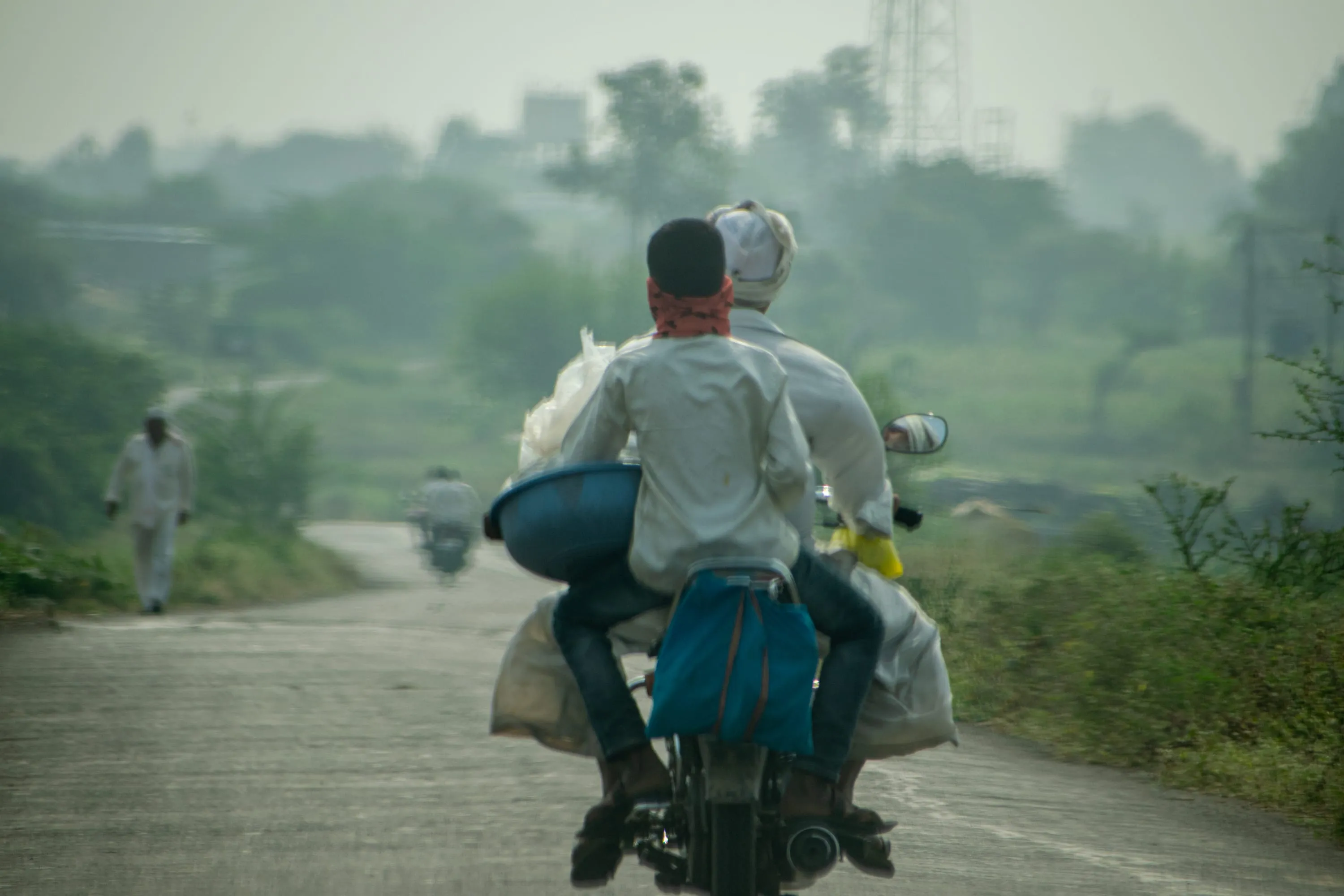 Two men riding a loaded motorcycle away down a wet road in morning haze, a third walking ahead.