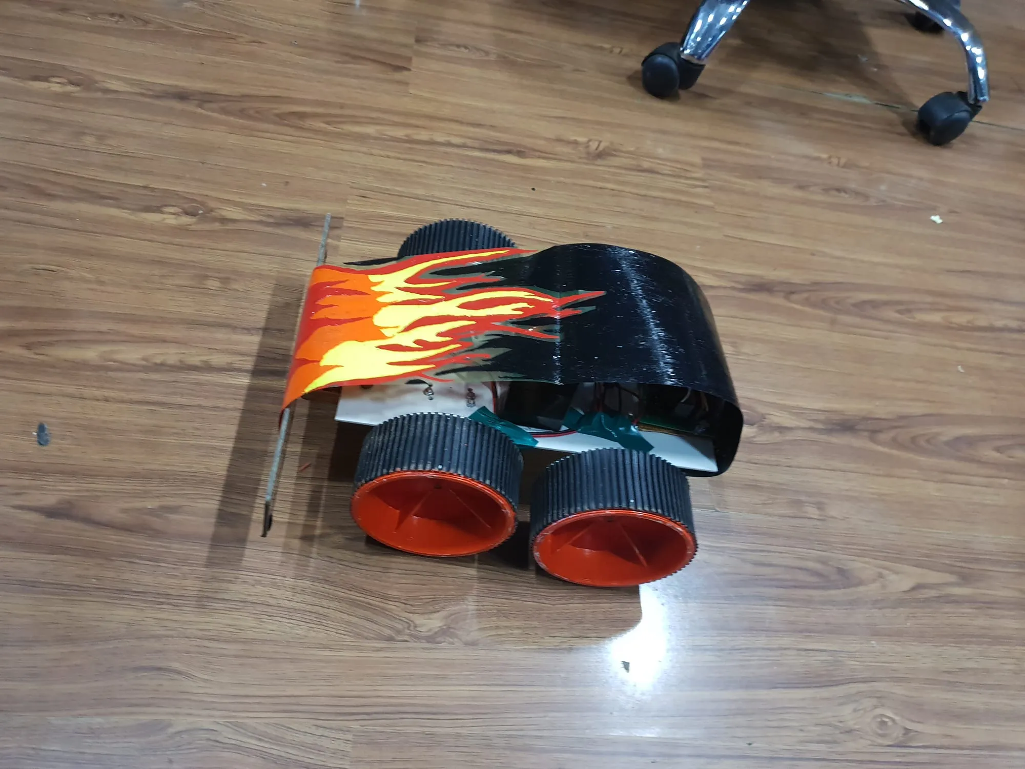 A small four-wheeled bot on a wooden floor: black ribbed tyres on orange hubs, a black shell hand-painted with orange and yellow flames, and a flat white front wing.